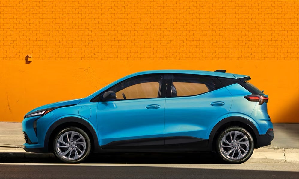 A bright blue 2027 Chevrolet Bolt electric SUV parked in front of an orange brick wall